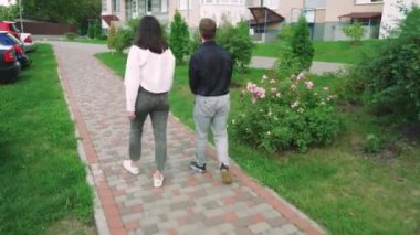 Bir kadın ve genç bir adam, sıradan giysiler içinde, kolları omzuna sarılı bir şekilde bir yerleşim tesisinin yakınlarında dolaşarak, şehirde rahat bir atmosfer yaratıyor.