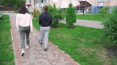 Bir kadın ve genç bir adam, sıradan giysiler içinde, kolları omzuna sarılı bir şekilde bir yerleşim tesisinin yakınlarında dolaşarak, şehirde rahat bir atmosfer yaratıyor.