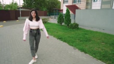 Genç bir kadın, pembe ceketli, modern bir apartmanın yanında güvenle yürüyor. Yaşam tarzı ve kentsel yaşam temaları için mükemmel bir sahne