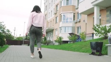 Omzunun üzerinden pembe ceketli bir kadın modern bir yerleşim yerinin yanında yürüyor, rahat bir şehir yaşam tarzını gösteriyor.