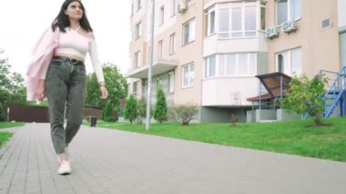 Omzunun üzerinden pembe ceketli bir kadın modern bir yerleşim yerinin yanında yürüyor, rahat bir şehir yaşam tarzını gösteriyor.