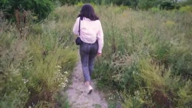 Omuzlarında siyah bir çanta taşıyan genç bir kadın, açık yeşil ve vahşi bitkilerle çevrili bir patika boyunca yürüyor. Yaşam tarzı ve açık hava sahneleri için mükemmel.