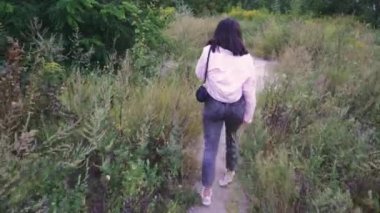 Omuzlarında siyah bir çanta taşıyan genç bir kadın, açık yeşil ve vahşi bitkilerle çevrili bir patika boyunca yürüyor. Yaşam tarzı ve açık hava sahneleri için mükemmel.