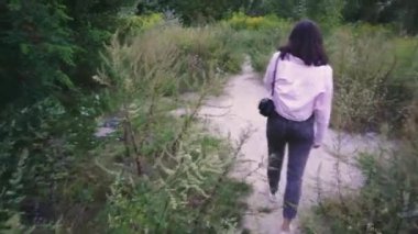 Omuzlarında siyah bir çanta taşıyan genç bir kadın, açık yeşil ve vahşi bitkilerle çevrili bir patika boyunca yürüyor. Yaşam tarzı ve açık hava sahneleri için mükemmel.