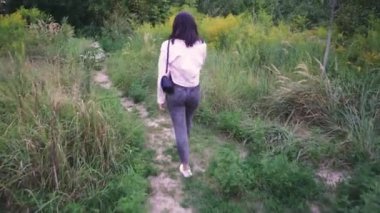 Omuzlarında siyah bir çanta taşıyan genç bir kadın, açık yeşil ve vahşi bitkilerle çevrili bir patika boyunca yürüyor. Yaşam tarzı ve açık hava sahneleri için mükemmel.