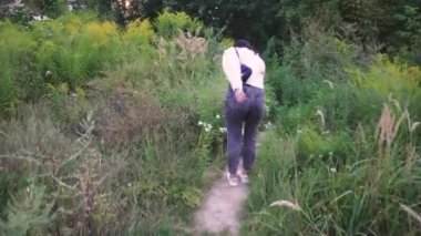 Omuzlarında siyah bir çanta taşıyan genç bir kadın, açık yeşil ve vahşi bitkilerle çevrili bir patika boyunca yürüyor. Yaşam tarzı ve açık hava sahneleri için mükemmel.