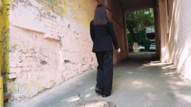 Bir kemer yolunda emin adımlarla ilerleyen, siyah takım elbiseli bir kadın, modern karmaşıklığı aktaran ve tamir edilmesi ve yer değiştirmesi gereken bir evin cephesi tarafından çerçevelenir.