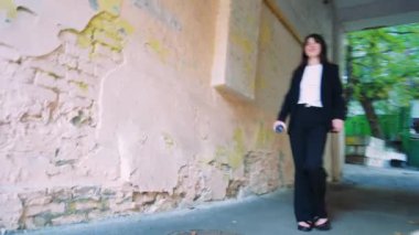 Bir kemer yolunda emin adımlarla ilerleyen, siyah takım elbiseli bir kadın, modern karmaşıklığı aktaran ve tamir edilmesi ve yer değiştirmesi gereken bir evin cephesi tarafından çerçevelenir.