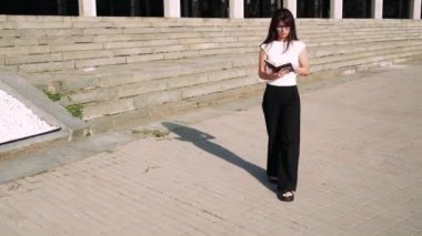 Siyah saçlı bir kız, büyük adımlarla zarif bir şekilde yürüyor. Arka planda büyük bir bina var. Siyah pantolon ve beyaz tişört giymiş.