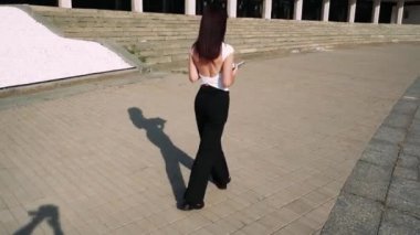 Siyah saçlı bir kız, büyük adımlarla zarif bir şekilde yürüyor. Arka planda büyük bir bina var. Siyah pantolon ve beyaz tişört giymiş.