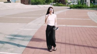 Bu video siyah pantolonlu ve beyaz tişörtlü bir kızı, arkasında sütunları olan görkemli bir bina tarafından çerçevelenmiş, etkileyici adımların yanından geçerken görüntülüyor.