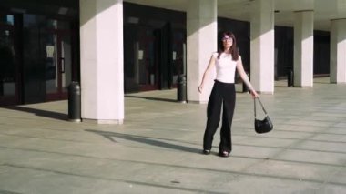 Resimli bir ortamda, siyah pantolonlu ve beyaz tişörtlü bir kız merdivenlerde büyük sütunları olan bir binanın yanında yürüyor ve sahneye zarafet katıyor.