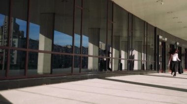 Siyah saçlı, siyah ceketli bir kız yerden tavana cam duvarları olan modern bir binanın yanında fayanslı zeminde koşuyor. Siyah pantolon ve beyaz tişört giymiş.