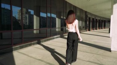 Siyah saçlı, siyah ceketli bir kız yerden tavana cam duvarları olan modern bir binanın yanında fayanslı zeminde koşuyor. Siyah pantolon ve beyaz tişört giymiş.