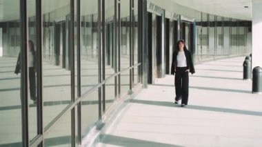 Siyah saçlı genç bir kız, modern cam duvarlı bir binanın yanında siyah bir ceket taşıyarak fayanslarla koşuyor. Siyah pantolon ve beyaz tişört giyiyor.