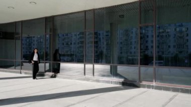 Siyah saçlı genç bir kız, modern cam duvarlı bir binanın yanında siyah bir ceket taşıyarak fayanslarla koşuyor. Siyah pantolon ve beyaz tişört giyiyor.