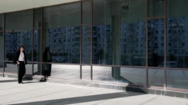 Siyah saçlı genç bir kız, modern cam duvarlı bir binanın yanında siyah bir ceket taşıyarak fayanslarla koşuyor. Siyah pantolon ve beyaz tişört giyiyor.