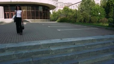 Kafasında güneş gözlüğü olan sıradan giyinmiş bir kız beton merdivenlerde elinde debriyaj ve laptop çantası, beyaz tişört ve siyah pantolon giyerek yürüyor.