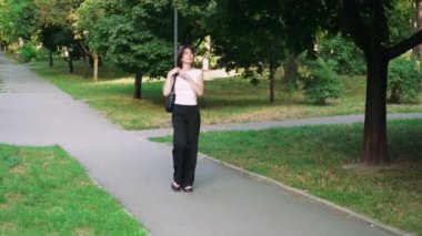 Kafasında gözlük olan siyah saçlı bir kız parkta günün tadını çıkarıyor. Kız parkın arka sokağında yürüyor ve elinde siyah bir çanta taşıyor.