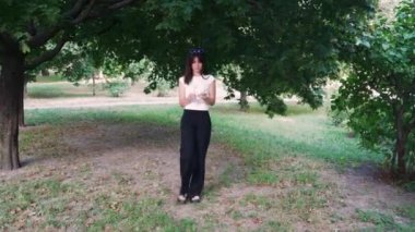 Parkta yürürken, siyah saçlı ve güneş gözlüklü bir kız siyah pantolon ve açık renk bir atlet giyiyor.