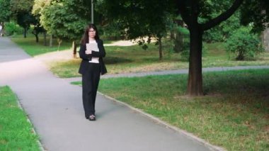 Gözlüklü bir kız yeşil bir parkta yürürken elinde bir dizüstü bilgisayar tutuyor. Klasik siyah takım elbiseli bir kadın avukat ya da ofis çalışanı gibi görünür.