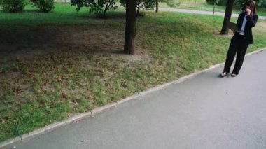 Beyaz tişörtlü ve siyah takım elbiseli bir kız elinde dizüstü bilgisayarla parkta yürüyor ve siyah saçlarında güneş gözlüğü var.