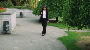 Siyah saçlı şık bir kız siyah bir çantayla parkta yürüyor, siyah klasik bir takım elbise giyiyor, avukat ya da iş kadını gibi görünüyor.