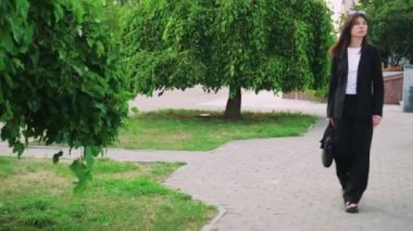 Siyah saçlı şık bir kız siyah bir çantayla parkta yürüyor, siyah klasik bir takım elbise giyiyor, avukat ya da iş kadını gibi görünüyor.