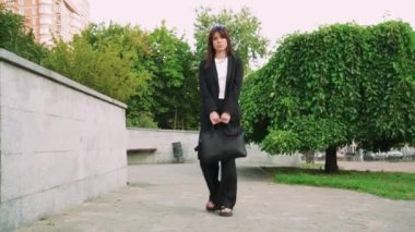 Siyah saçlı şık bir kız siyah bir çantayla parkta yürüyor, siyah klasik bir takım elbise giyiyor, avukat ya da iş kadını gibi görünüyor.