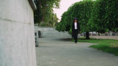 Beyaz tişörtlü ve siyah pantolonlu sıradan giyinmiş bir kadın arka planda evler ve yeşilliklerle dolu bir parkta geziniyor ve tasasız bir sahne yaratıyor.