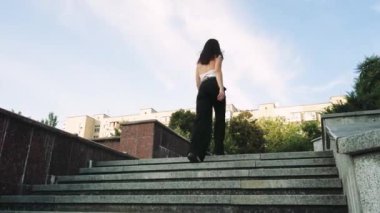 Beyaz atletli ve siyah pantolonlu genç bir kadın arka planda evler ve yeşil ağaçlarla beton basamaklardan aşağı yürüyor.
