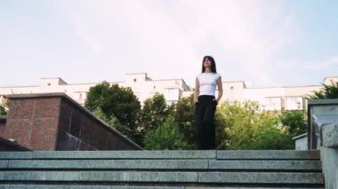 Beyaz atletli ve siyah pantolonlu genç bir kadın arka planda evler ve yeşil ağaçlarla beton basamaklardan aşağı yürüyor.