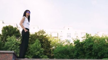 Beyaz atletli ve siyah pantolonlu bir kadın granit levha boyunca yürür, arkasında evler ve ağaçlar görülebilir. Gündüz vakti yaz parkı