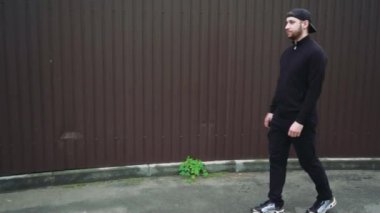 Siyah ceketli ve şapkalı bir adam arka planda bordo bir metal çitle sessiz bir caddede yürüyor. Sokak tarzı, kentleşme kavramı ile birleştirilmiştir.