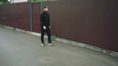 Siyah ceketli ve şapkalı bir adam arka planda bordo bir metal çitle sessiz bir caddede yürüyor. Sokak tarzı, kentleşme kavramı ile birleştirilmiştir.