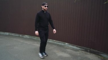 Siyah ceketli ve şapkalı bir adam arka planda bordo bir metal çitle sessiz bir caddede yürüyor. Sokak tarzı, kentleşme kavramı ile birleştirilmiştir.