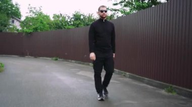 Siyah giysili genç bir adam kaldırımlı bir cadde boyunca güneş gözlüğü ve beyzbol şapkası takarak güvenle yürüyor. Kentsel sokak tarzı