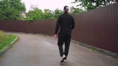 Siyah giysili genç bir adam kaldırımlı bir cadde boyunca güneş gözlüğü ve beyzbol şapkası takarak güvenle yürüyor. Kentsel sokak tarzı