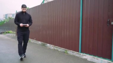 Siyahlı bir adam elinde kitapla şehir caddesinde yürüyor. Önünde kahverengi metal bir çit ve modern binalar var. Kentsel yaşam tarzı, modern yaşam ve gelişigüzel anlar için mükemmel