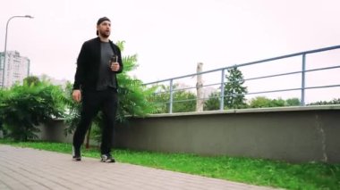 Siyah kıyafetli genç bir adam elinde içkiyle şehir kaldırımında yürüyor, çevresi yeşillik ve binalarla çevrili. Kentsel yaşam tarzı ve günlük rutin kavramlar için mükemmel
