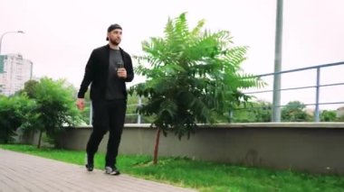 Siyah kıyafetli genç bir adam elinde içkiyle şehir kaldırımında yürüyor, çevresi yeşillik ve binalarla çevrili. Kentsel yaşam tarzı ve günlük rutin kavramlar için mükemmel
