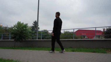 Siyah kıyafetli genç bir adam elinde içkiyle şehir kaldırımında yürüyor, çevresi yeşillik ve binalarla çevrili. Kentsel yaşam tarzı ve günlük rutin kavramlar için mükemmel