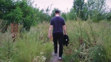 Siyah eşofmanlı bir adam vahşi bitkiler ve ağaçlarla çevrili doğa yürüyüşü patikalarını keşfediyor. Doğa, macera ve açık hava videoları için uygun