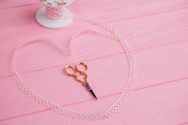 Lace ribbon, elegantly wrapped in the shape of a heart on a pink background, and in the middle lie metal scissors that add charm to DIY projects and crafts clipart