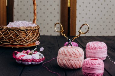Çeşitli el aletleri ahşap bir sepete istiflenir. Koyu renkli bir masada pembe iplikler, iğneler ve makas yastığı var.