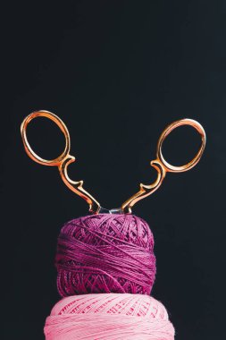 Yarn in skeins of pink and purple shades are stacked on top of each other on a black background, golden scissors are stuck into these skeins clipart