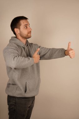 Tamam işareti. Yakışıklı, kendine güvenen, gri kapüşonlu bir adam baş parmağını tek başına kaldırıyor. Gri arka planda sakallı yakışıklı genç bir adam iyi niyet gösterisi yapıyor. Yan görünüm