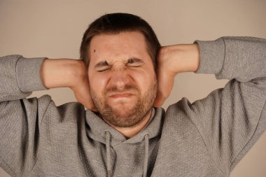 Genç bir adamın portresi elleriyle kulaklarını kapatıyor ve gözlerini gri renkte kapalı tutuyor. Umutsuz, sinirli, sakallı bir adamın fotoğrafı. Ellerini başının üstünde tutuyor ve gözlerini kapatıyor. Depresyona giriyor.