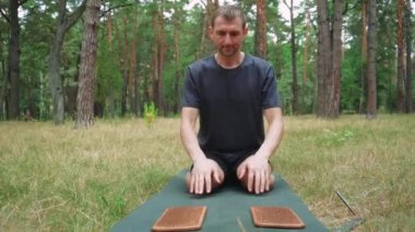 Bir adam, ormanın ortasında, temiz havada, yeşil bir minder üzerinde iğneli tahtaların yardımıyla yoga egzersizleri yapıyor.