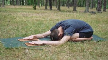 Bir adam, ormanın ortasında, temiz havada, yeşil bir minder üzerinde iğneli tahtaların yardımıyla yoga egzersizleri yapıyor.